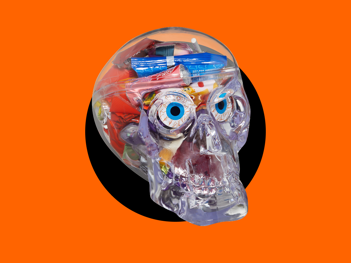 Crânio de plástico transparente cheio de doces de Halloween, incluindo olhos de doce e embalagens de doces variados, sobre um fundo laranja e preto.
