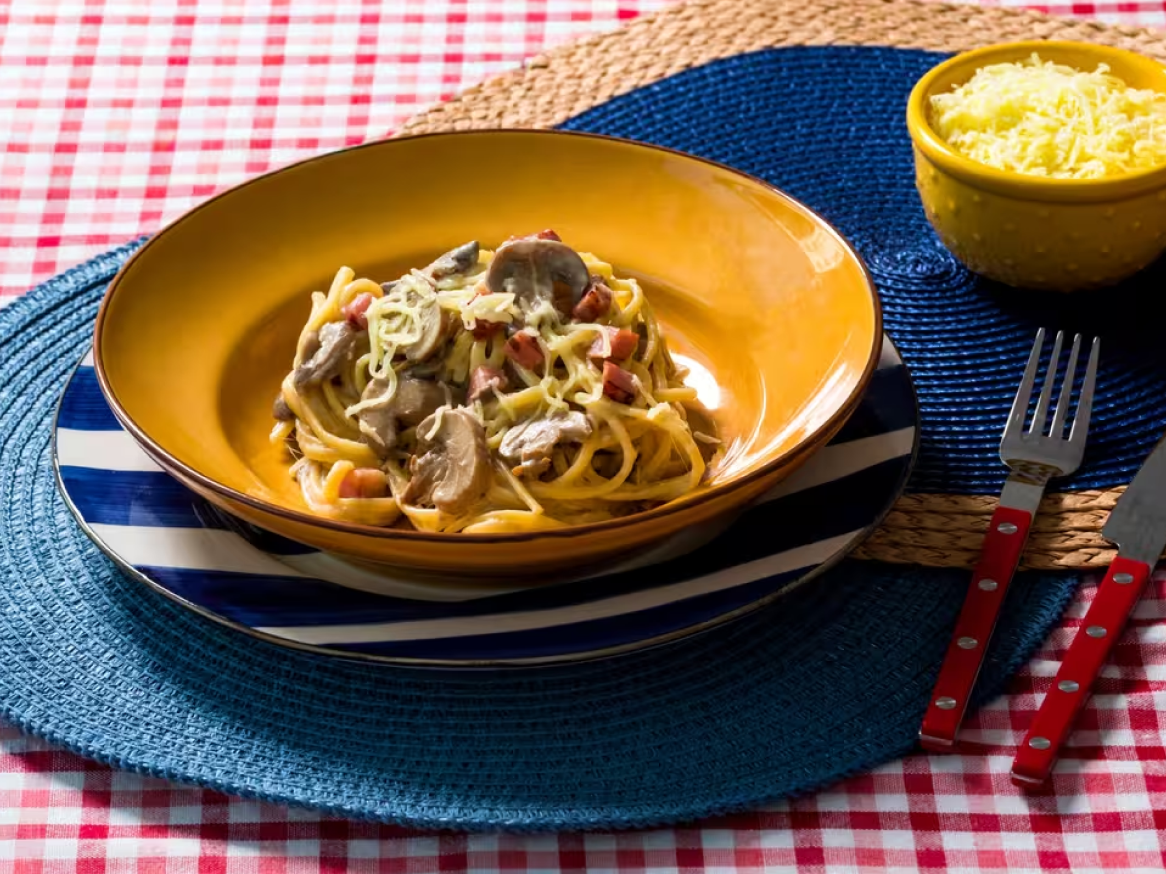 Esparguete com cogumelos e bacon num prato amarelo, com queijo ralado e talheres.