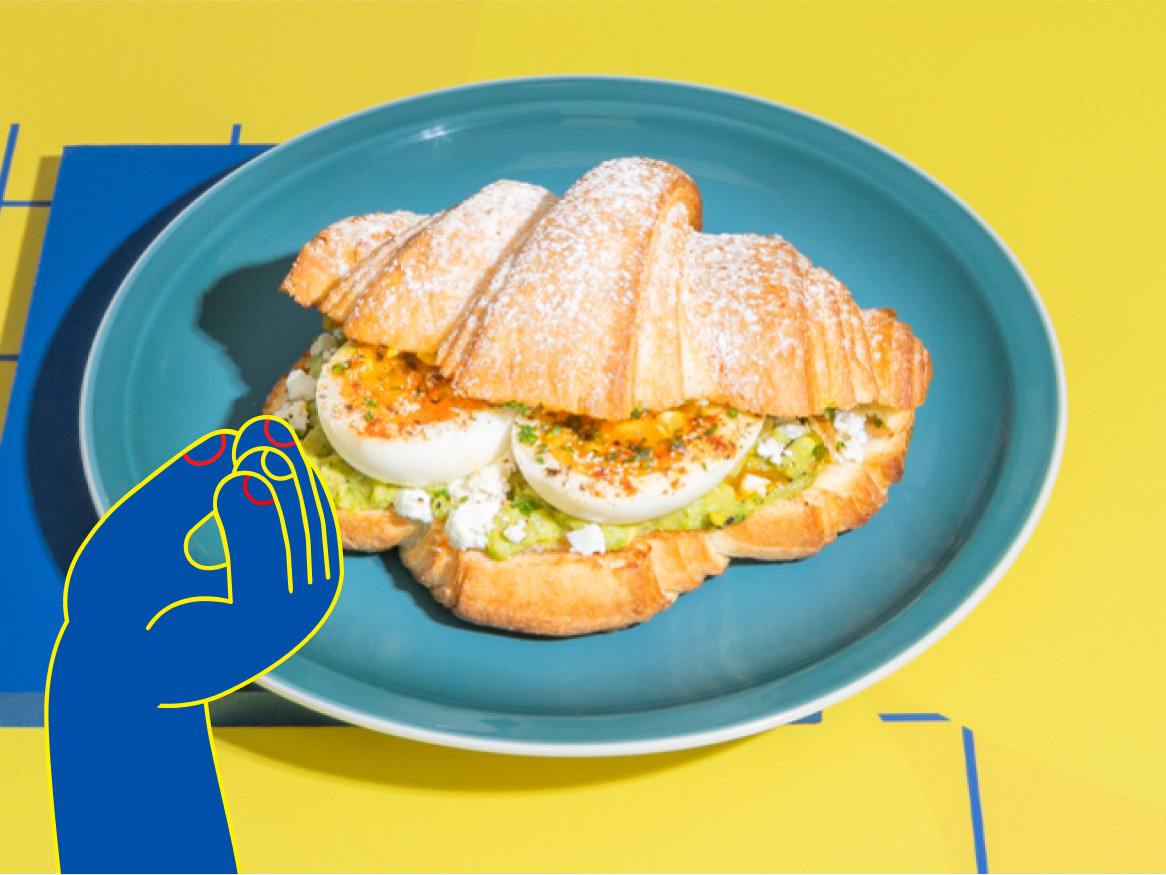 Croissant com ovos cozidos, abacate e queijo feta num prato azul.