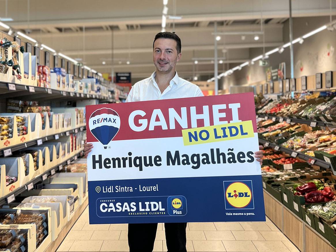 Um homem sorridente segura um cartaz 'GANHEI NO LIDL' num supermercado, com produtos de mercearia ao fundo.