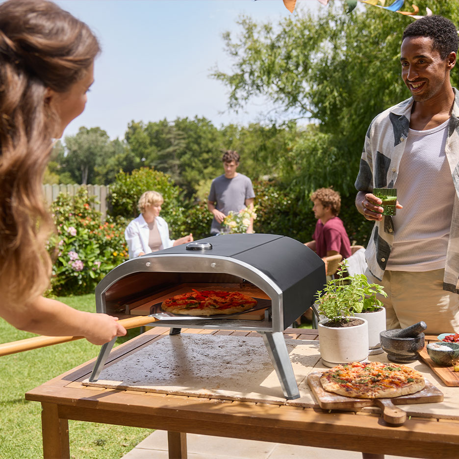 Família numa festa no jardim, mulher a colocar pizza num forno, homem a segurar uma bebida.